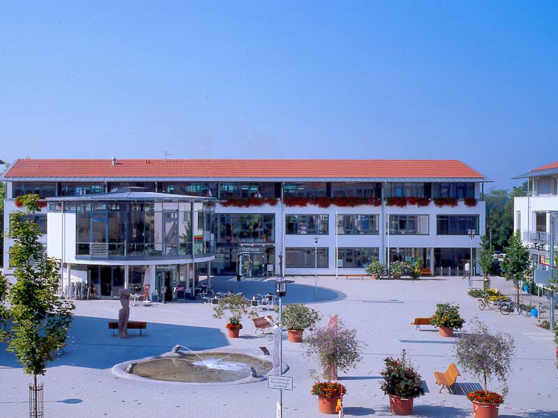 Eine moderne Gebäudestruktur mit rotem Ziegeldach umgibt einen offenen Platz, der mit Bäumen, Blumenbeeten und einem kleinen Brunnen gestaltet ist.