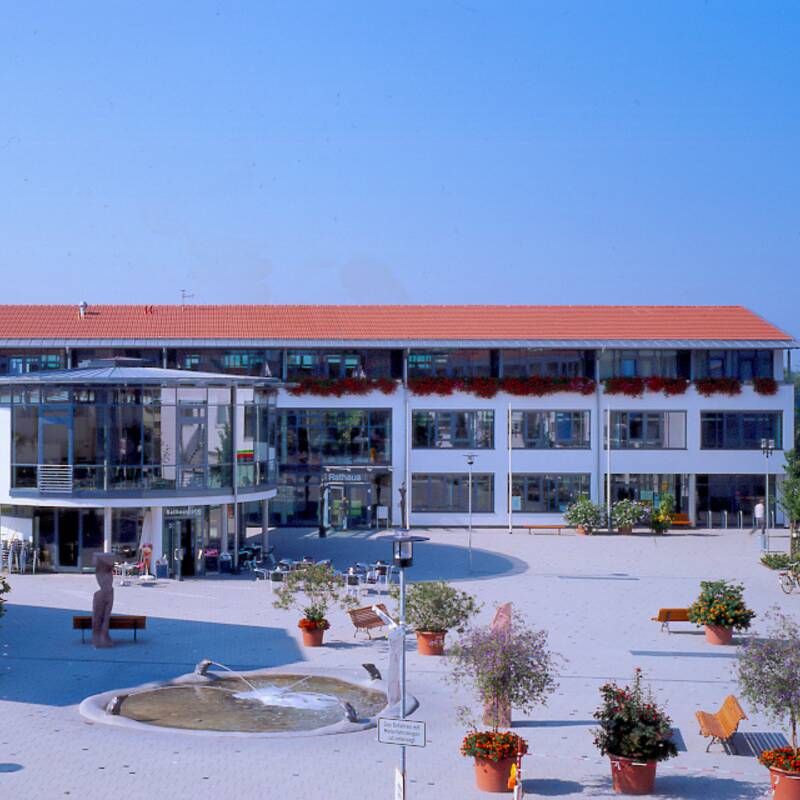 Eine moderne Gebäudestruktur mit rotem Ziegeldach umgibt einen offenen Platz, der mit Bäumen, Blumenbeeten und einem kleinen Brunnen gestaltet ist.