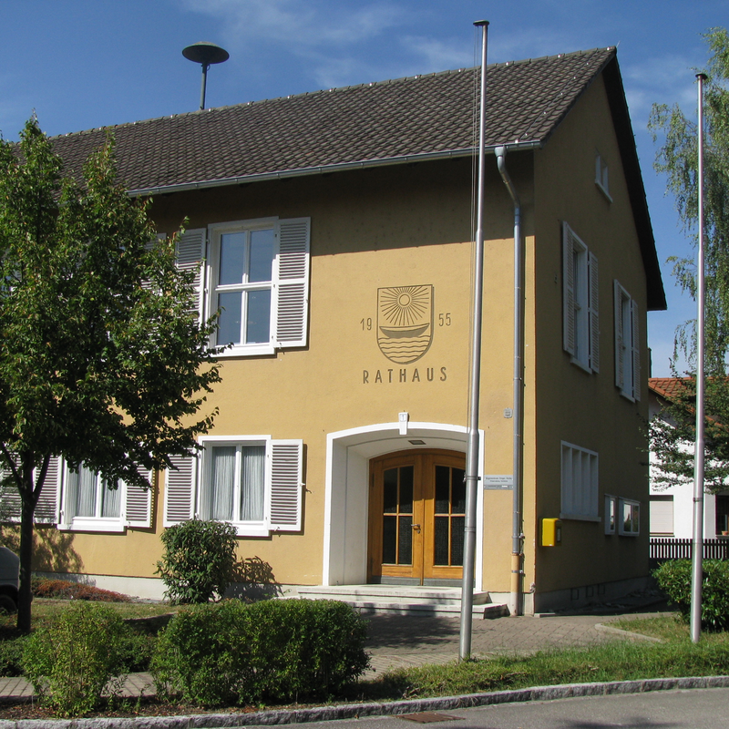 Ein zweistöckiges Gebäude mit gelber Fassade und dem Schriftzug "Rathaus", umgeben von Bäumen und Büschen, steht unter klarem Himmel.