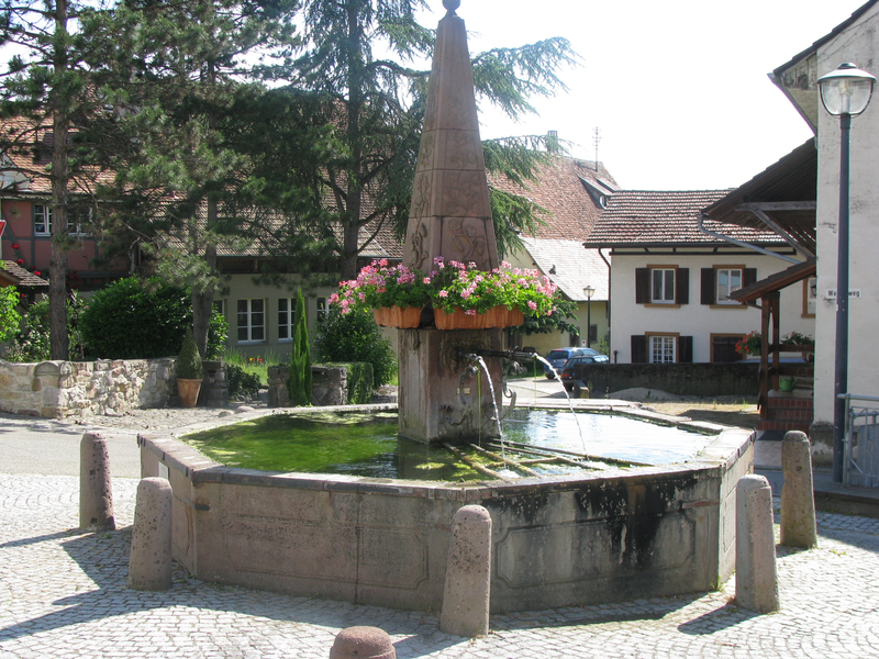 Ein achteckiger Steinbrunnen mit einem Obelisken in der Mitte steht in einer gepflasterten Umgebung, umgeben von Gebäuden, mit sprudelndem Wasser und blühenden Blumen an der Spitze.