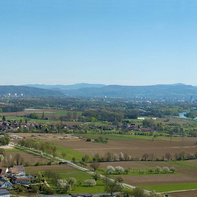 Die Panoramaaufnahme zeigt eine weite ländliche Landschaft mit Feldern und Wäldern im Vordergrund und einer Siedlung im Hintergrund, unter einem klaren blauen Himmel.