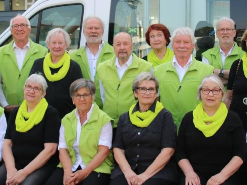 Eine Gruppe älterer Menschen trägt weiße Hemden und grüne Westen oder Tücher und posiert vor einem weißen Bus.