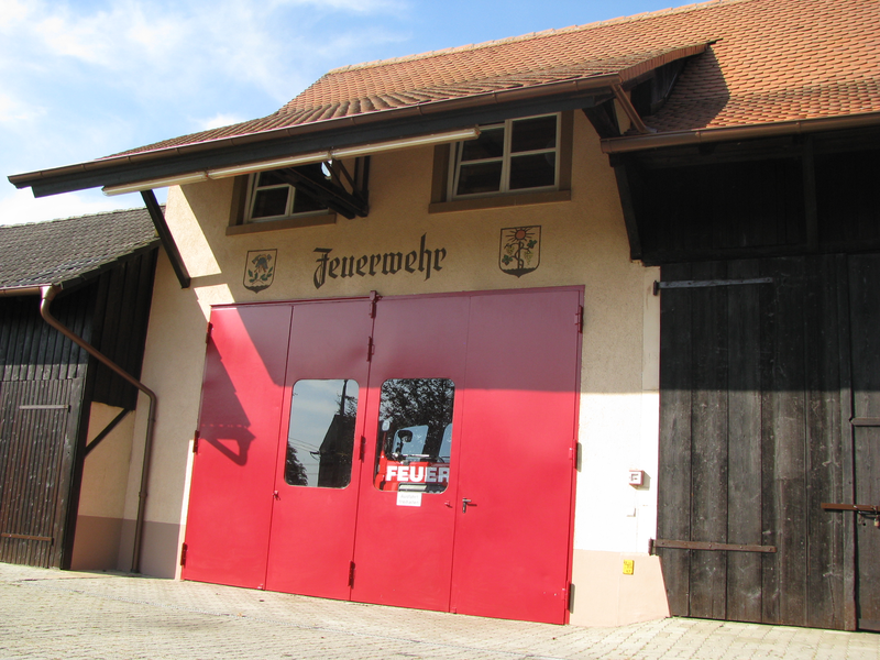 Ein Gebäude mit roten Garagentoren und der Aufschrift "Feuerwehr" über dem Eingang ist unter einem klaren blauen Himmel zu sehen.