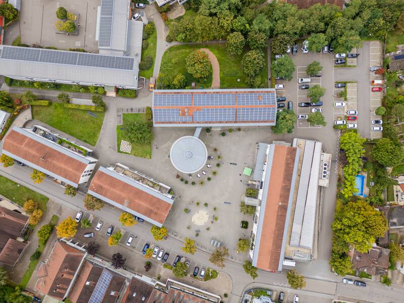 Die Luftaufnahme zeigt einen Gebäudekomplex mit mehreren rechteckigen Gebäuden, einem runden Hof, Solarpaneelen auf den Dächern und parkenden Autos, umgeben von Bäumen und Straßen.