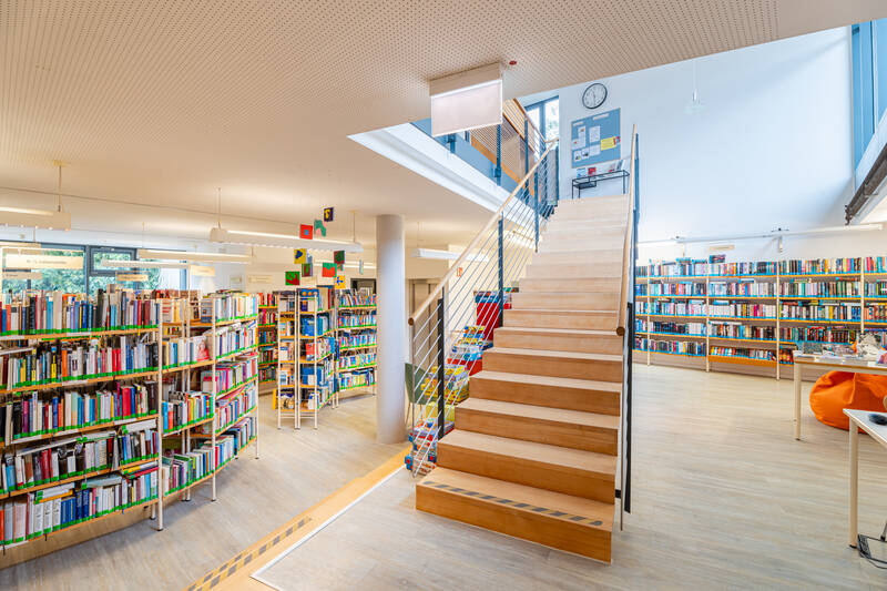 Ein helles, modernes Bibliotheksinterieur mit Bücherregalen, einer Holztreppe und bequemen Sitzgelegenheiten.