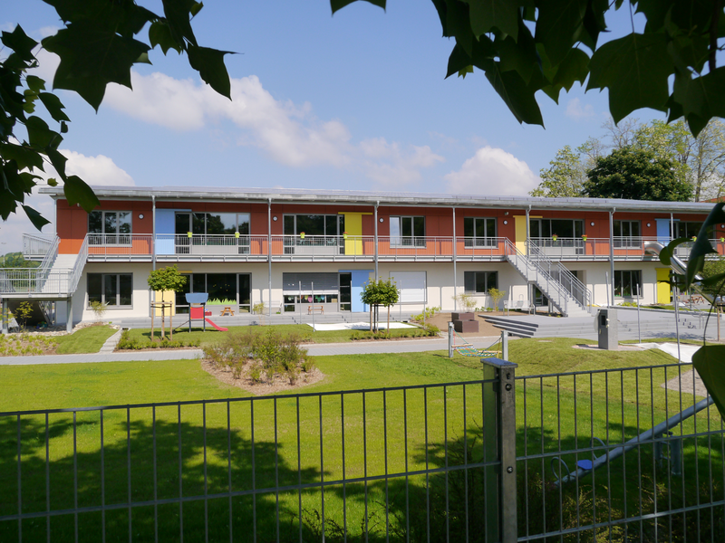 Ein zweistöckiges Gebäude mit farbigen Wänden und Fluchttreppen steht hinter einem grünen, gepflegten Garten mit Spielplatz, umrahmt von Bäumen.