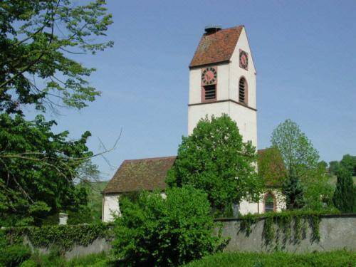Auf dem Bild ist eine Kirche mit einem Glockenturm und rotem Ziegeldach zu sehen, die von üppigem Grün umgeben ist.