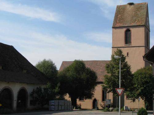 Das Bild zeigt eine historische Kirche mit einem hohen Turm und einem dreieckigen Dach, umgeben von Bäumen, auf einem sonnigen Tag.