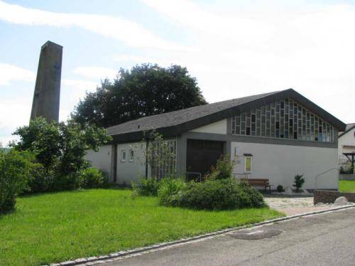 Ein modernes Gebäude mit schrägem Dach steht in einer grünen Umgebung, flankiert von einem hohen, schmalen Turm und Bäumen.