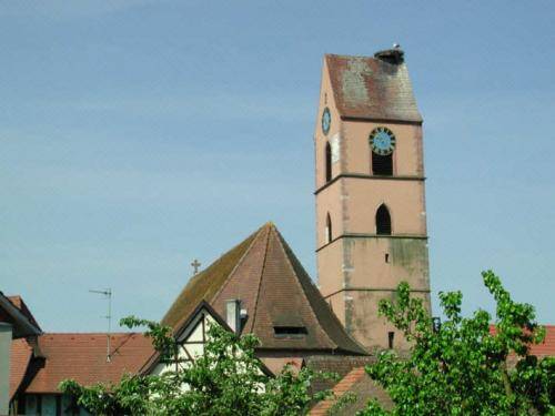 Ein alter Kirchturm mit einer Uhr ist von roten Dächern und grünen Bäumen umgeben.