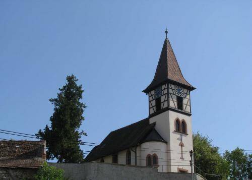 Das Bild zeigt eine Kirche mit einem Fachwerkturm und einem spitzen Dach vor einem klaren blauen Himmel.