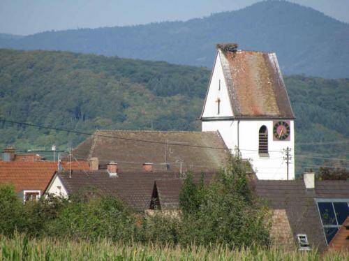 Ein historisches Kirchturm mit Ziegeldach und Uhr steht über den Dächern eines Dorfes, umgeben von grünen Bäumen und bewaldeten Hügeln im Hintergrund.