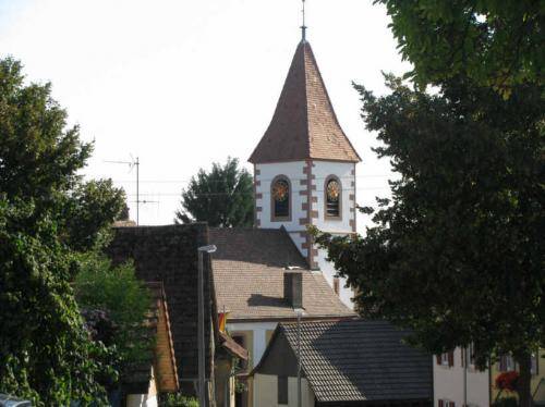 Eine malerische Dorfansicht mit einem Kirchturm im Vordergrund, umgeben von grünen Bäumen und traditionellen Gebäuden.