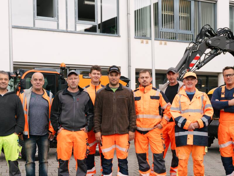 Eine Gruppe von neun Personen in Arbeitskleidung posiert vor einem Gebäude neben einer Baumaschine.