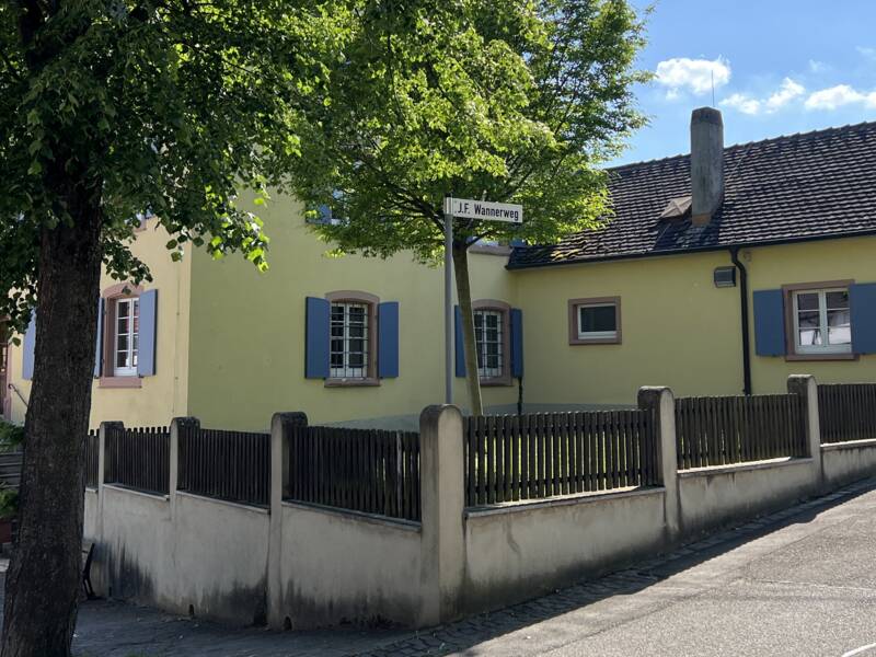 Ein gelbes Haus mit blauen Fensterläden steht unter einem großen Baum, umgeben von einem Holzzaun und einem gepflasterten Weg.
