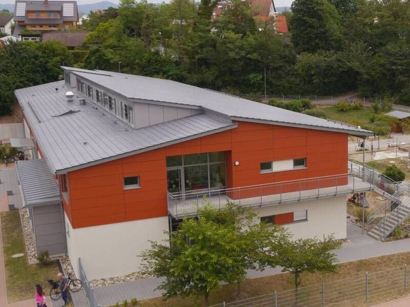 Ein modernes Gebäude mit roter und weißer Fassade steht neben einem umzäunten Park, in dem Menschen auf Grünflächen spazieren gehen und spielen, im Hintergrund sind Bäume und weitere Gebäude zu sehen.