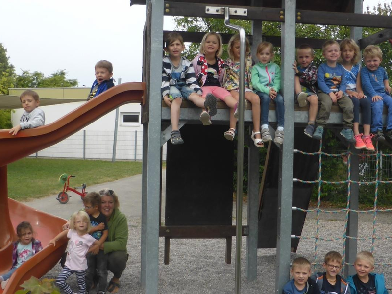 Eine Gruppe von Kindern sitzt und spielt auf einem Klettergerüst und einer Rutsche draußen auf einem Spielplatz, begleitet von zwei Erwachsenen.