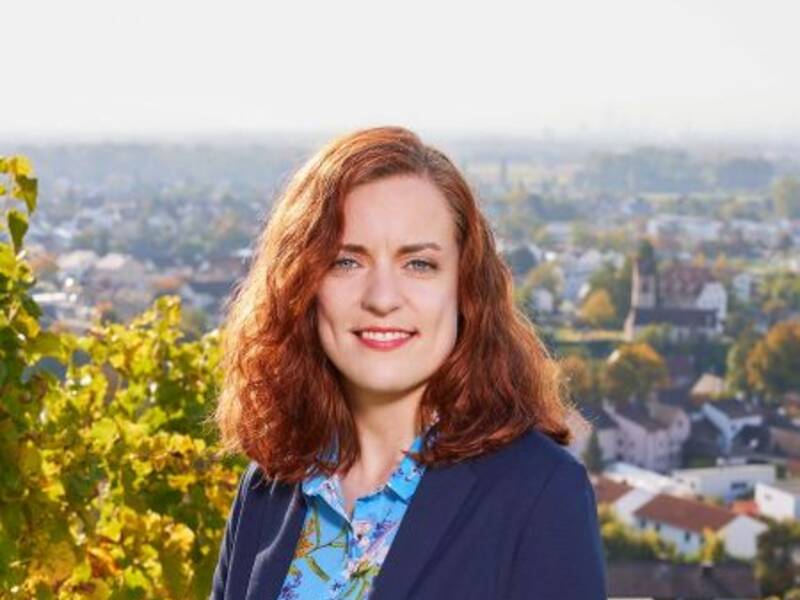 Eine Person mit lockigem, rötlichem Haar steht lächelnd in einem Weinberg mit einem Dorf im Hintergrund.