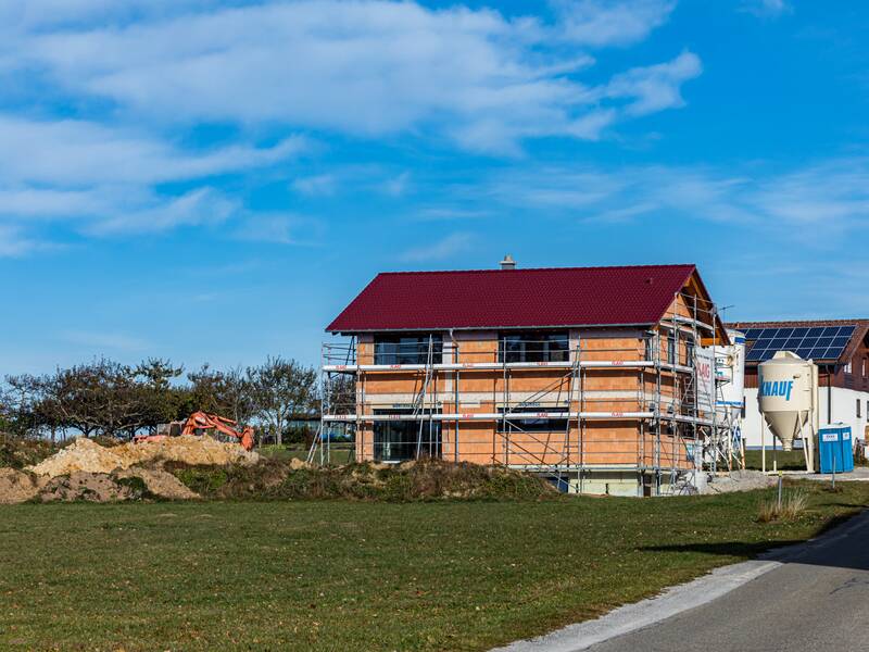 Ein neu gebautes Haus mit Gerüst steht auf einer Wiese, umgeben von Baugeräten und einem blauen Himmel im Hintergrund.